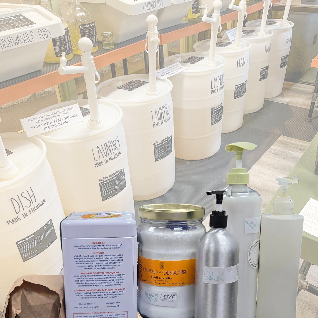A collection of handmade laundry and cleaning products displayed on a table, including large white plastic canisters with manual pumps, a silver pump bottle, a clear bottle with a green pump, a jar with a gold lid, and a light purple box, set against a background of more similar containers.