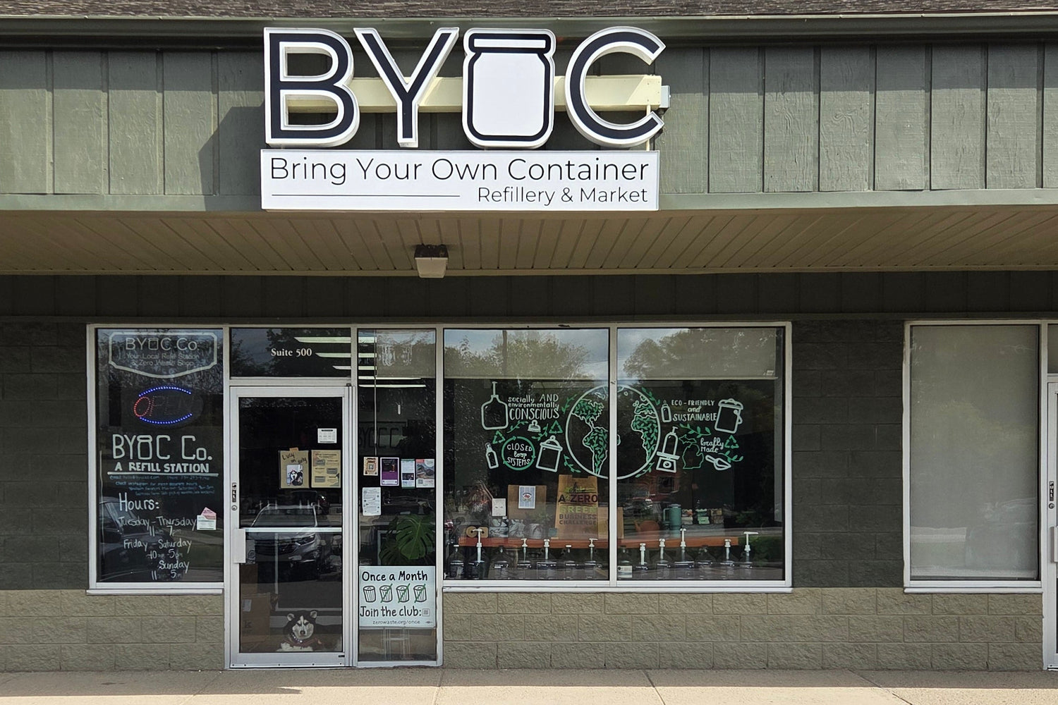 BYOC (Bring Your Own Container) dispensary storefront with a gray exterior, large glass windows, and a cloudy sky above, with a paved parking lot in the foreground.