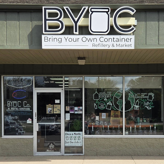 BYOC (Bring Your Own Container) dispensary storefront with a gray exterior, large glass windows, and a cloudy sky above, with a paved parking lot in the foreground.